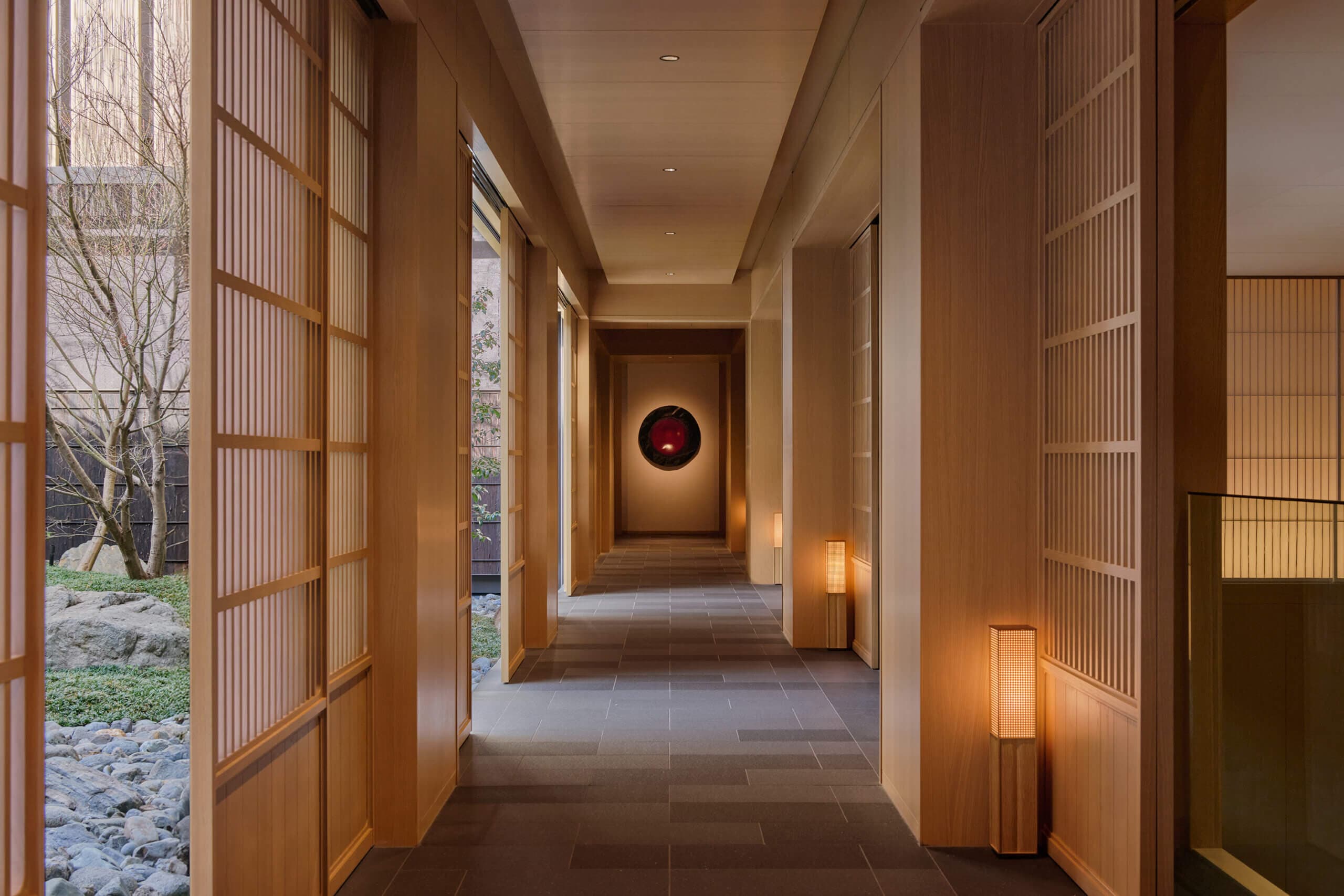 Long wood-lined arrival corridor at Capella Kyoto with shoji screens, courtyard garden glimpses, and a circular red artwork at the far wall